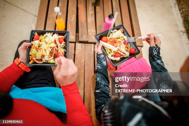 two athletes using plastic eating utensils to eat a healthy protein salad from a plastic take-out box while sitting together on a park bench - plastic cutlery stock pictures, royalty-free photos & images