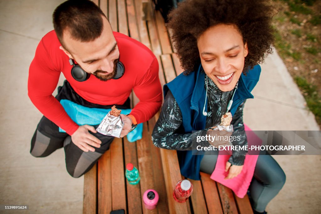 Young active woman eating a protein bar with her partner while sitting on a bench in a city park