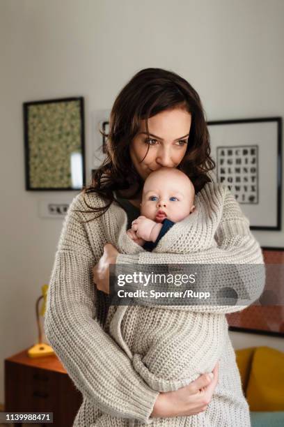 woman kissing baby son, wrapping him in her cardigan - mom holding baby stock-fotos und bilder