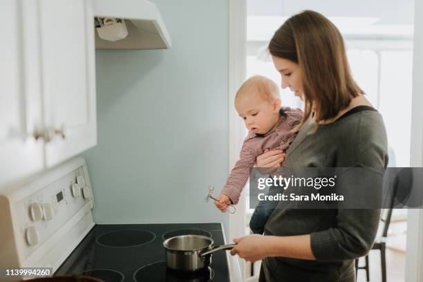 mother carrying baby son while cooking at hob - burner stove top stockfoto's en -beelden