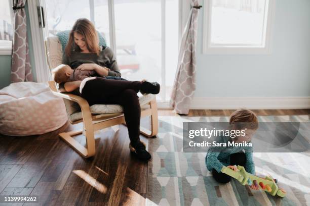 female toddler playing in living room while mother breastfeeding baby son - two kids playing with hose stock-fotos und bilder