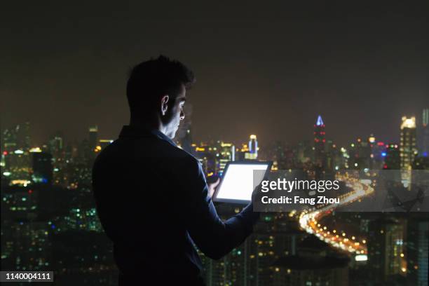 silhouetted young businessman looking at digital tablet in front of skyscraper office window at night, shanghai, china - night before fotografías e imágenes de stock