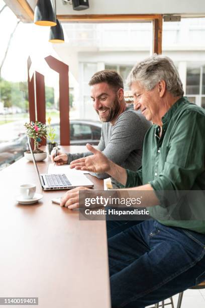 cheerful latin american men at a bakery in a meeting looking at laptop while talking and smiling - coffee meeting stock pictures, royalty-free photos & images