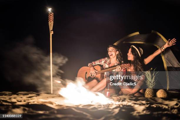 zwei glückliche freundinnen campieren an einem strand neben dem feuer. - brand on guitar stock-fotos und bilder