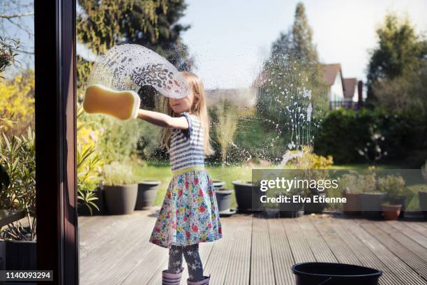 child (6-7) washing patio window with sponge and soapy water, outside on a sunny day - girls in wet dresses stock-fotos und bilder