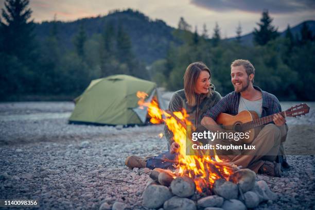 young couple sitting by campfire playing guitar - brand on guitar stock-fotos und bilder