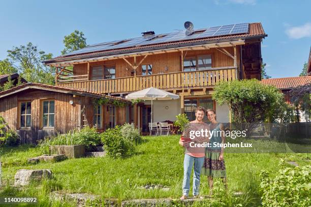 portrait of young couple, holding pot plant, standing in front of house with solar panelled roof - bavarian man in front of house stockfoto's en -beelden