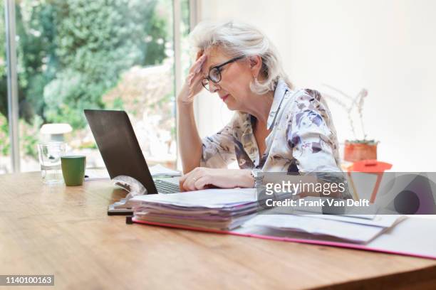 senior woman sitting at table, using laptop, worried expression - frau aktenordner stock-fotos und bilder