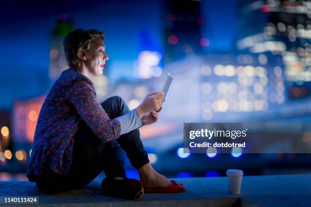 mature businesswoman sitting on waterfront using  digital tablet at night, london, uk - night before fotografías e imágenes de stock