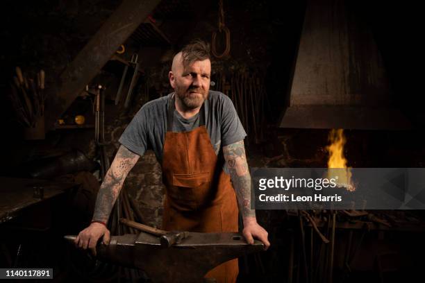 mature male blacksmith leaning against anvil in workshop, portrait - forzudo fotografías e imágenes de stock
