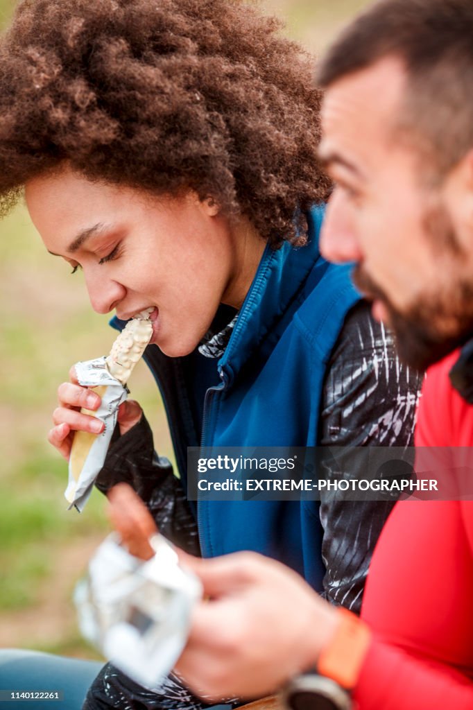 Young active woman eating a protein bar with her partner while sitting on a bench in a city park
