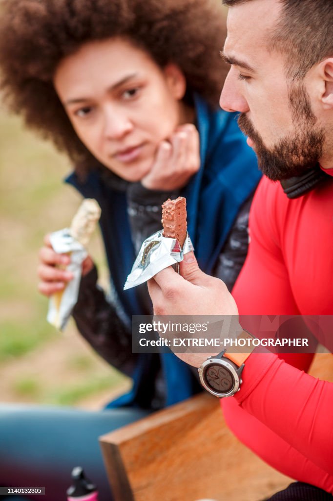 Junge Sportlerin genießt mit ihrem Partner in einer Proteinbar, während sie auf einer Bank in einem Stadtpark sitzt