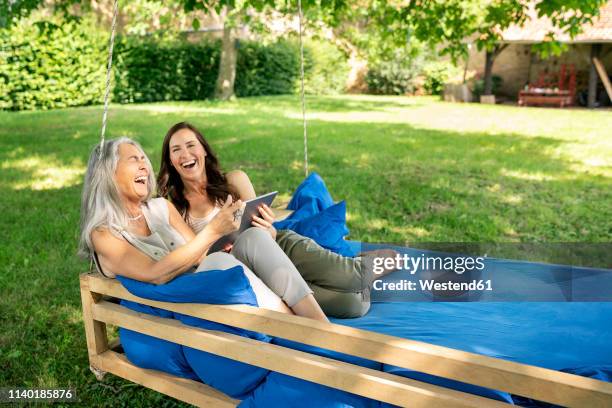 two laughing women relaxing on a hanging bed in garden using tablet - garden of laughs stockfoto's en -beelden