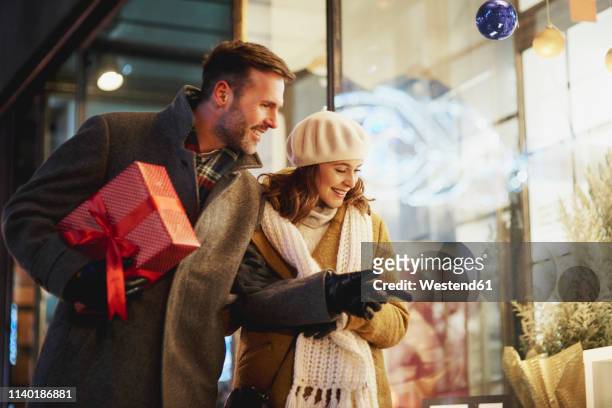 smiling couple looking in shop window at christmas time - christmas shopping stock-fotos und bilder