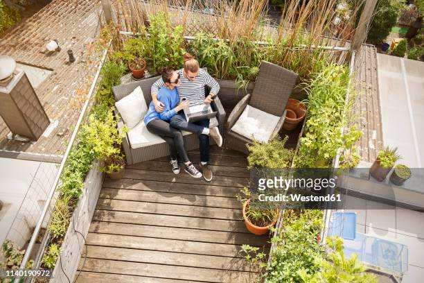 young couple relaxing on their balcony, sitting on couch, using laptop - couple balcony stock-fotos und bilder