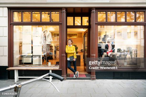 young woman standing in door of a fashion store, holding laptop - store window stock pictures, royalty-free photos & images