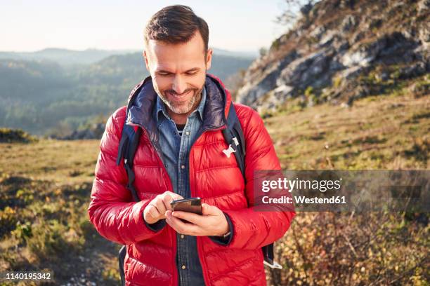 smiling man on a hiking trip in the mountains checking cell phone - mann auf berg mit smartphone stock-fotos und bilder