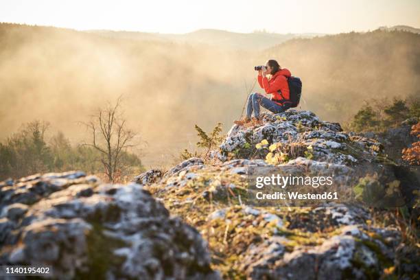 woman on a hiking trip in the mountains sitting on rock looking through binoculars - fernglas stock-fotos und bilder