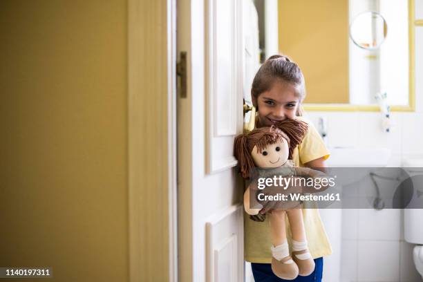 portrait of smiling little girl standing in doorframe at home holding a doll - puppe stock-fotos und bilder