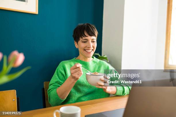 happy short-haired woman with green pullover working with laptop in home office eating cornflakes in a bowl - eating cereal stock pictures, royalty-free photos & images