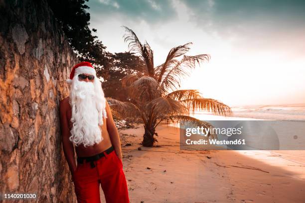 thailand, man dressed up as santa claus leaning against wall on the beach at sunset - santa leaning stock pictures, royalty-free photos & images