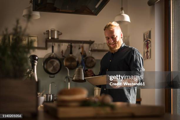 young man standing in kitchen, reading online recipe on his digital tablet - keukengereedschap stockfoto's en -beelden