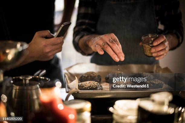 young man taking pictures of friend,  sprinkling sesame on burger buns on a baking tray - kochen nahaufnahme stock-fotos und bilder