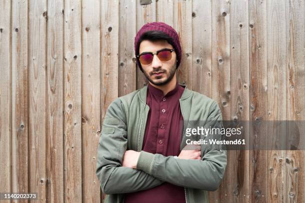 portrait of serious bearded man in purple knit hat and shirt with arms crossed wearing sunglasses over wooden background - arrogancia fotografías e imágenes de stock