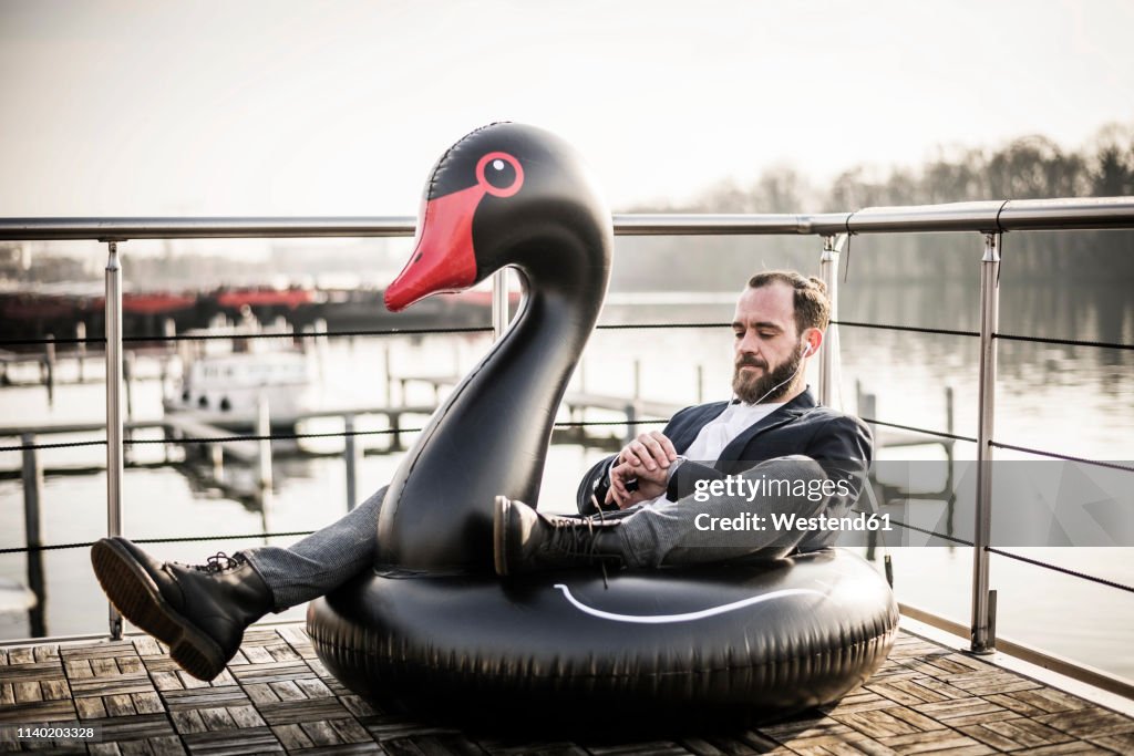 Hip businessman working on a houseboat, sitting a floating swan, using smartwatch