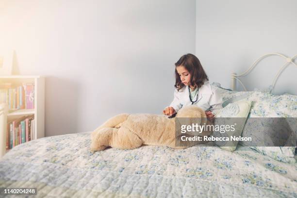 girl dressed up in lab coat sitting in bed using stethoscope on soft toy - labbett stock-fotos und bilder