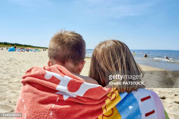 rear view of a girl and a boy covered in a towel and sitting together on a beach - wrapped in a towel stock-fotos und bilder