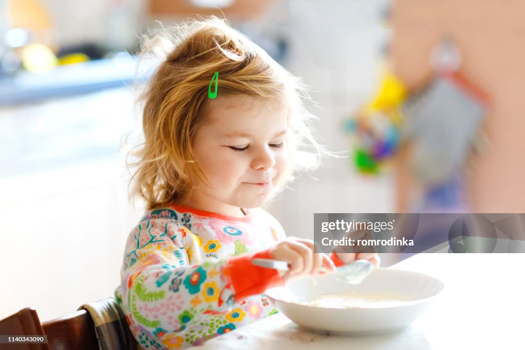 Adorable toddler girl eating healthy porridge from spoon for breakfast. Cute happy baby child in colorful pajamas sitting in kitchen and learning using spoon.