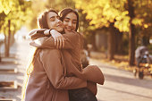 Side profile of young pretty smiling brunette twin girls hugging and having fun in casual coat standing close to each other at autumn sunny park alley on blurry background