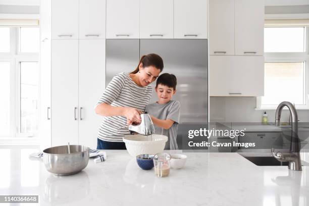 mother helping young son use a mixer while cooking in a modern kitchen - cocina electrodomésticos fotografías e imágenes de stock