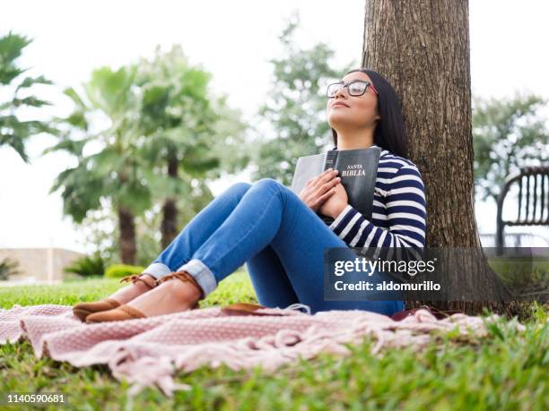 hübsche junge frau sitzt neben baum und hält bibel - women prayer stock-fotos und bilder