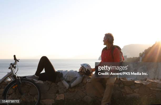 female biker relaxes on stone wall, with male companion - couché de soleil photos et images de collection