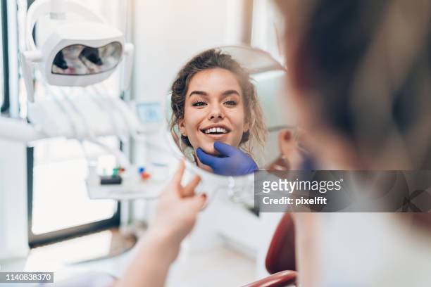 young woman looking in the mirror after a dental procedure - smiling dentist stock pictures, royalty-free photos & images