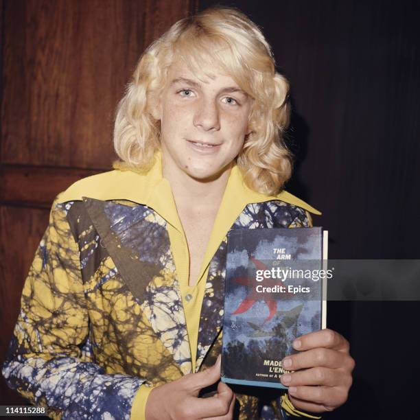 American actor and filmmaker Darby Hinton in a brightly coloured shirt poses with a copy of 'The Arm Of The Starfish' by Madeleine L'Engle, United...