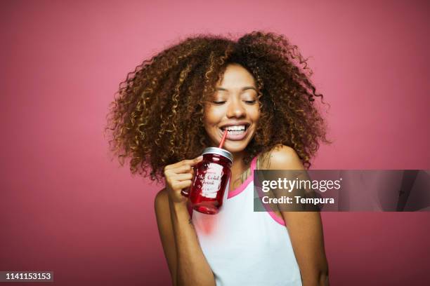 beautiful afro caribbean young woman in summer clothes against pink background. - woman drinking juice stock pictures, royalty-free photos & images