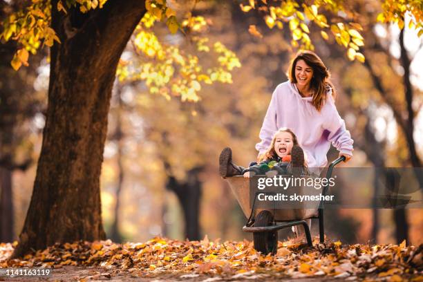 gelukkige moeder die haar kleine dochter in een wheelbarrow in het park duwt. - kruiwagen stockfoto's en -beelden