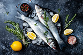 Fresh raw seabass fish on black stone background with spices, herbs, lemon. Culinary seafood background with ingredients for cooking. Top view