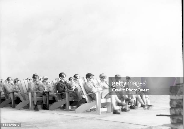 Various unidentified US military personnel, all wearing large googles, sit on Adirondack-style chairs as they witness a nuclear test, codenamed...
