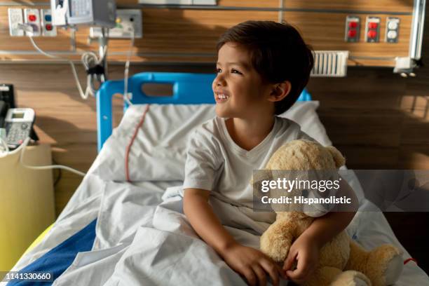 cute boy sitting down on hospital bed hugging his teddy bear smiling - stuffed toy stock pictures, royalty-free photos & images