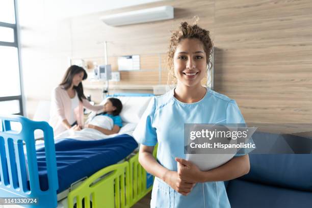 beautiful nurse facing camera smiling holding paperwork and little girl lying down on hospital bed while mom comforts her - woman smiling facing down stock pictures, royalty-free photos & images