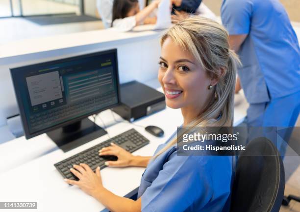 schöne freundliche krankenschwester an der rezeption des krankenhauses arbeitet am computer, während vor der kamera lächelnd - bürorezeption stock-fotos und bilder