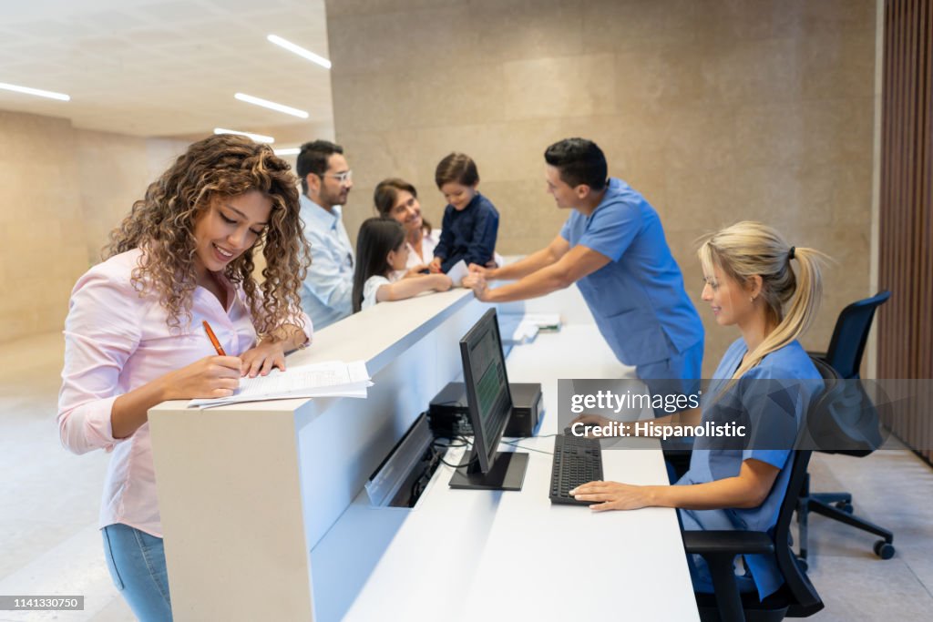 Beautiful latin american patient filling in a form at the hospital's front desk all smiling