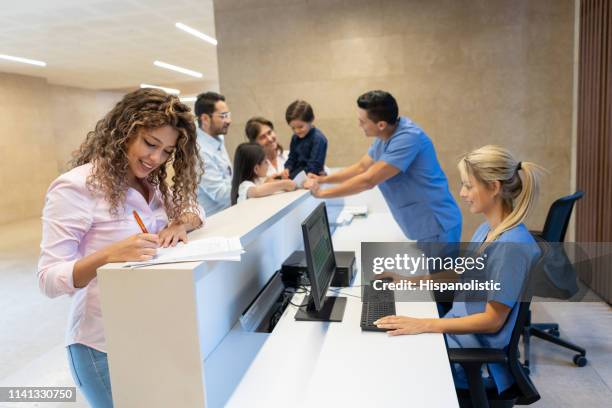 beautiful latin american patient filling in a form at the hospital's front desk all smiling - hospital reception stock pictures, royalty-free photos & images