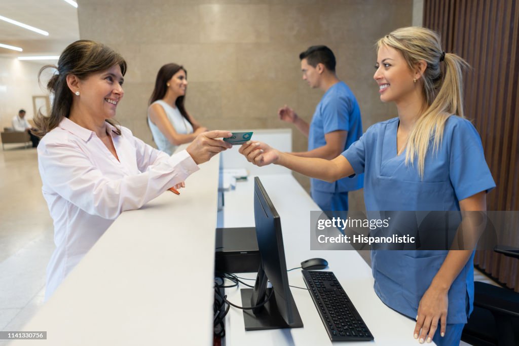 Mujer adulta madura entregando su tarjeta de seguro a la enfermera en la recepción del hospital tanto sonriendo
