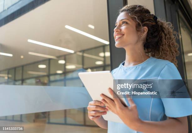 cheerful young beautiful nurse looking away through the window daydreaming and smiling while holding a tablet - nurse thinking stock pictures, royalty-free photos & images