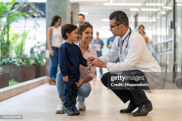 pediatra maschio che parla con il suo piccolo paziente che è in piedi accanto a sua madre tutti sorridenti - group of patients foto e immagini stock